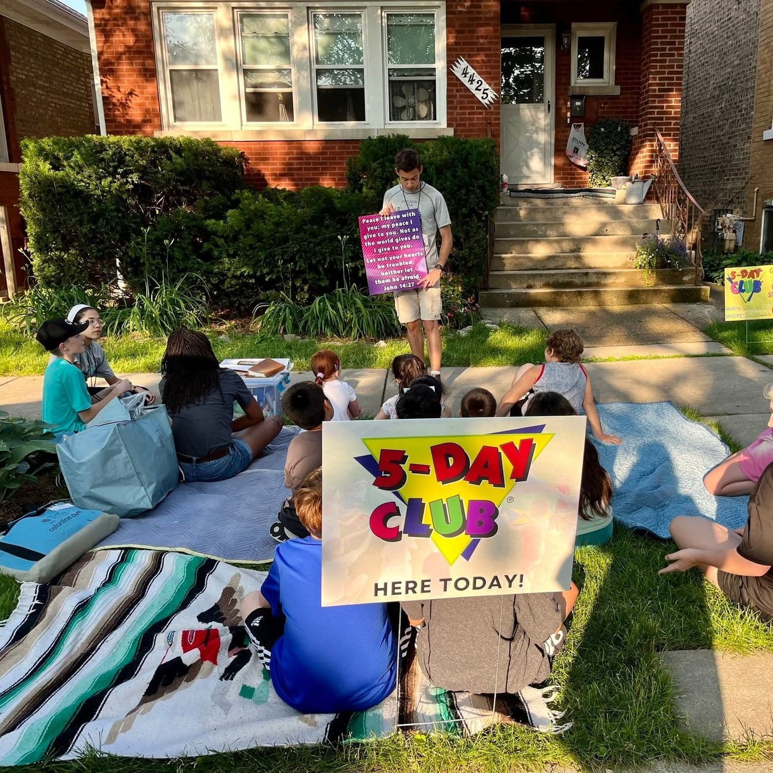 Children sit on blankets in a front yard behind a sign that reads 5-Day Club here today