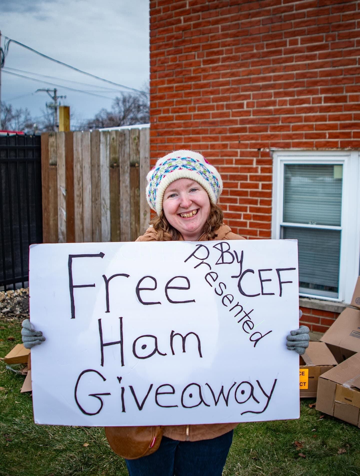 A smiling woman in a knit hat holds a hand-written sign that reads Free Ham Giveaway, presented by CEF