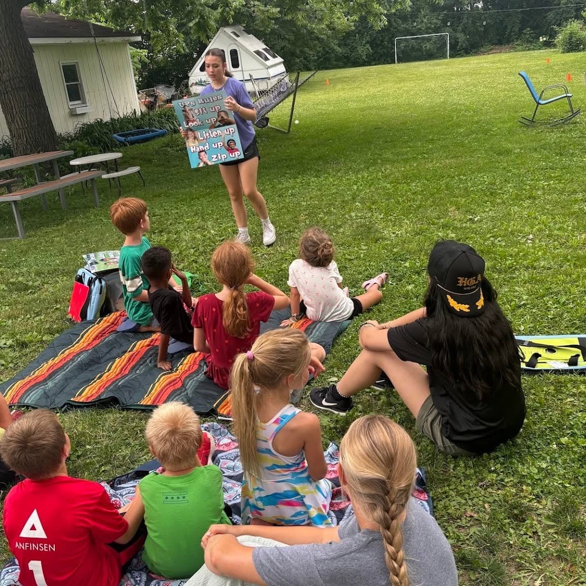Children sit on striped mats on the grass while a leader teaches