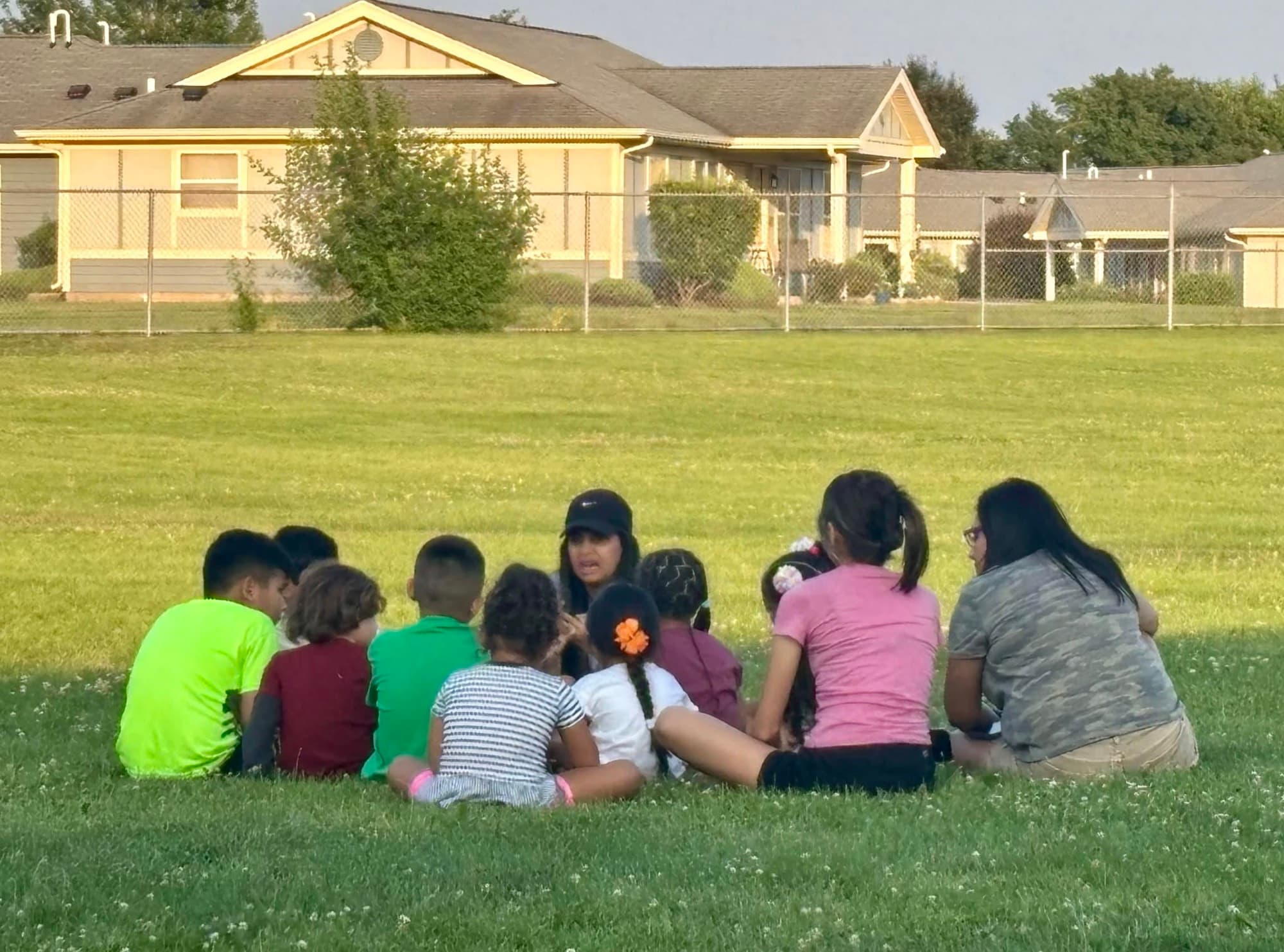A small group of children sit in a circle on a lawn in front of houses